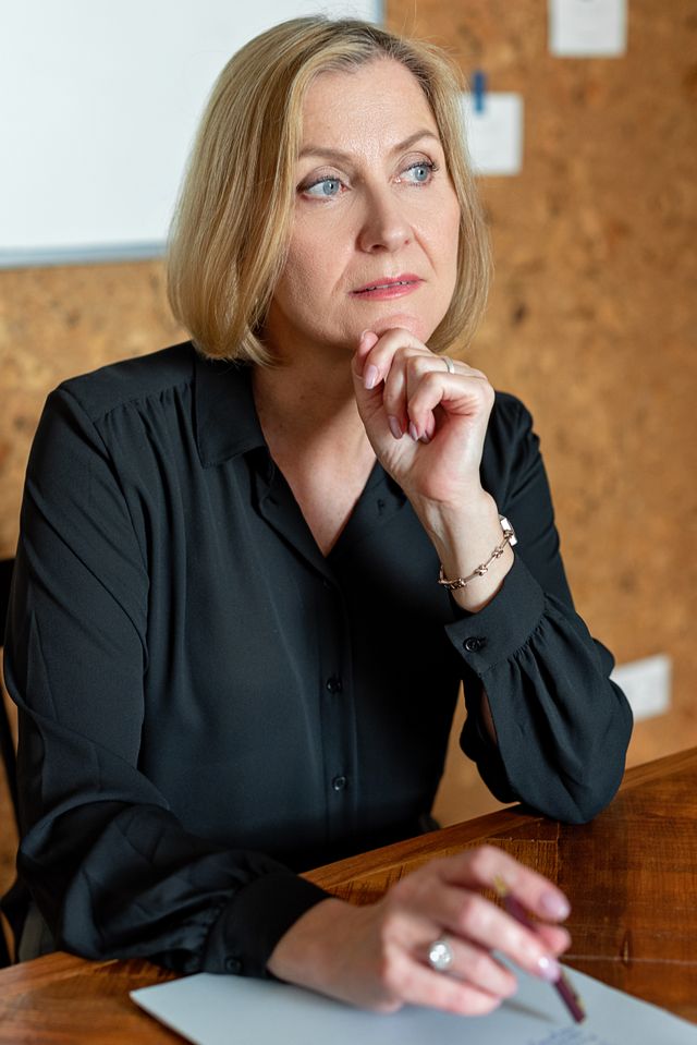 Mature businesswoman in black attire thinking at a wooden desk in a modern office.